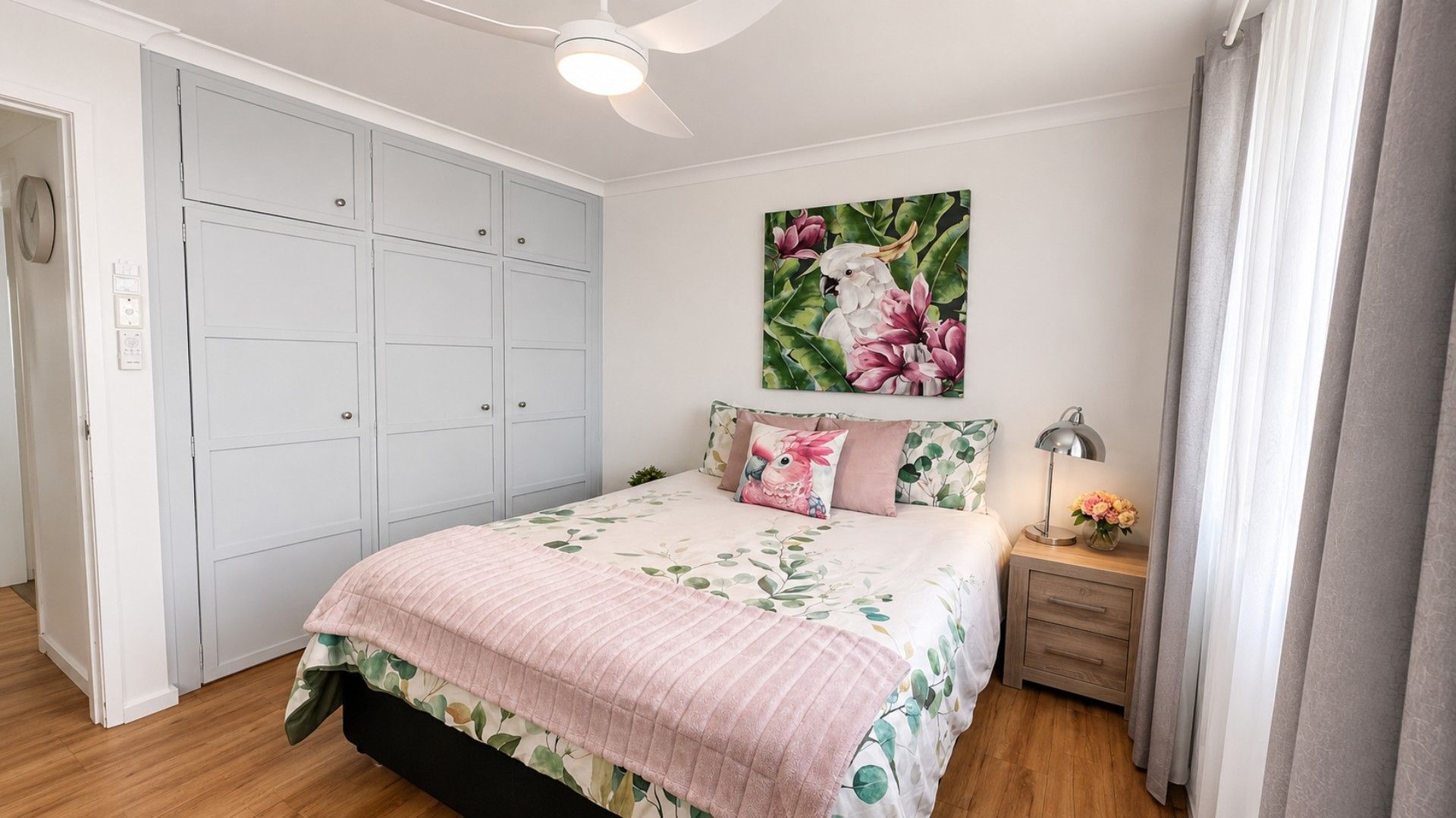 A bedroom with a bed, a nightstand, a ceiling fan, a wardrobe, and a floral painting on the wall.