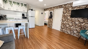 Una cocina moderna con armarios blancos, microondas integrado y refrigerador. Se observa una pared de piedra con un televisor montado y una silla color turquesa.