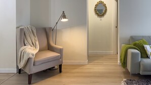 A living room with a grey armchair, a floor lamp, a sofa with a green throw, and a hallway leading to a door with a gold mirror. - Modern 2BR | Near Victoria Hospital (London)