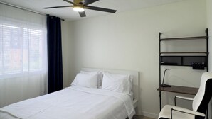 A hotel room with a bed, a desk, a chair, a ceiling fan, and a window with curtains. - Modern 2BR | Near Victoria Hospital (London)