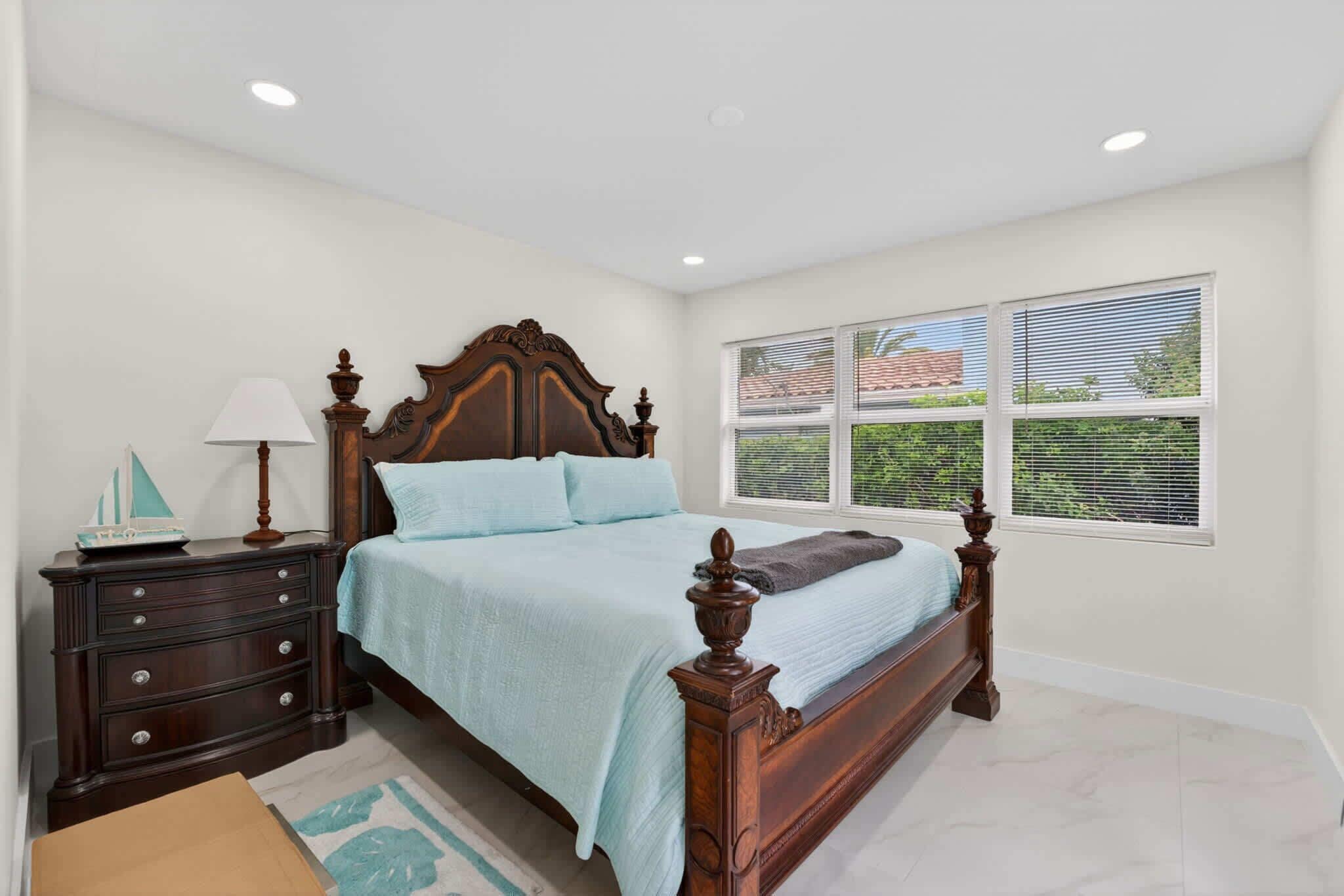A bedroom with a wooden bed, a nightstand, a lamp, and a window with blinds.