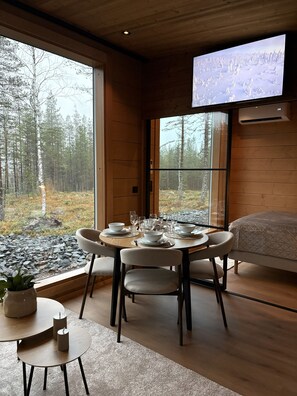 A modern interior with a dining area, a bed, and a large window offering a forest view.