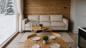 Un intérieur chaleureux avec un plafond en bois, un canapé, une table à manger dressée pour quatre personnes et une vue sur les arbres enneigés à l’extérieur.