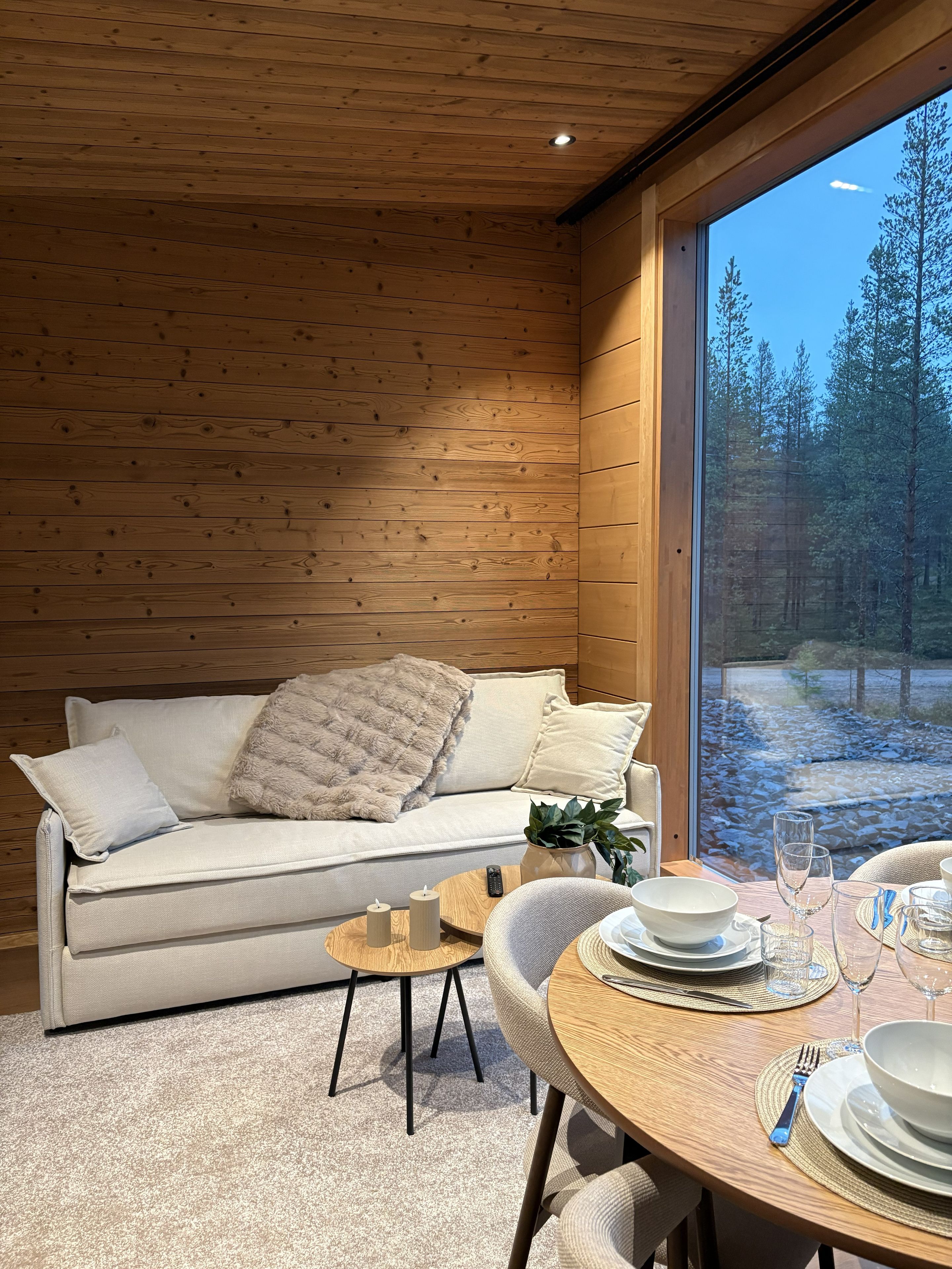 L’intérieur d’un chalet accueillant, avec un plafond en bois, un canapé agrémenté de coussins et une table à manger mise avec des assiettes, des verres et des couverts.