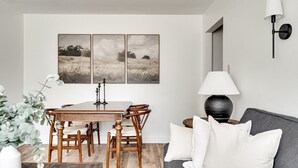 Un comedor con una mesa y sillas de madera, un sofá con cojines blancos, una lámpara de pared y cuadros en la pared.