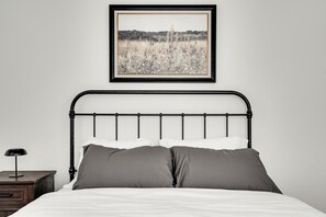 Ein Bett mit Metallgestell, weißer Bettwäsche und zwei grauen Kissen. Darüber hängt ein gerahmtes Bild.