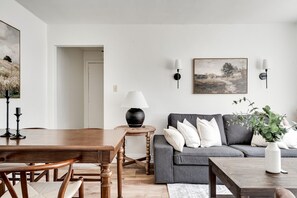 A living room with a wooden dining table, a grey sofa, and a small side table with a lamp. - Bright 2BR – Walk to Downtown Kingston & Lake! (Kingston)