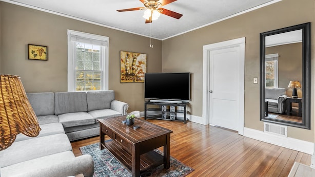 Un salon avec un ventilateur de plafond, un canapé gris, une table basse, un téléviseur et un grand miroir.