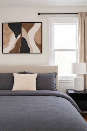 A bedroom with a bed, a nightstand, a lamp, a window with curtains, and a wall art piece. - Cozy 2BR – Walk to Downtown & University! (Kingston)