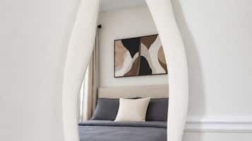Une chambre à coucher comprenant un lit, des oreillers, un tableau abstrait accroché au mur et un miroir d’une forme arquée originale.