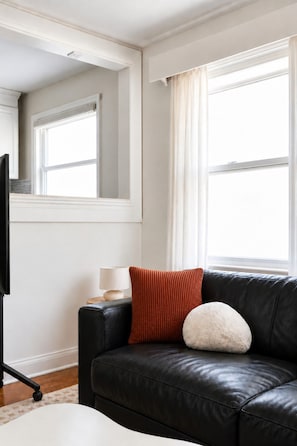 A modern living room with a black leather sofa, a white lamp, and a television on a stand. - Cozy 2BR – Walk to Downtown & University! (Kingston)