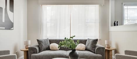 Una sala de estar con un sofá, dos mesas auxiliares, una mesa de centro y un ventanal con cortinas translúcidas.