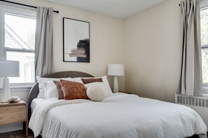 A neatly made bed with white bedding and brown pillows, a bedside table with a lamp, and a window with curtains. - Bright 1BR Near Queen’s, KGH & Waterfront! (Kingston)