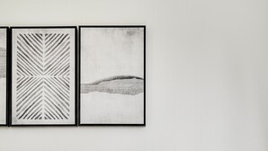 Ein ordentlich bezogenes Bett mit weißer Decke und zwei Kissen, ein Nachttisch mit Lampe sowie zwei gerahmte Kunstwerke darüber.