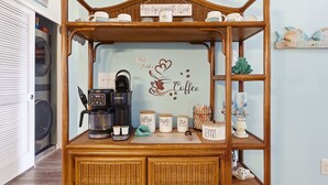 A coffee station with a sign reading 'But First... Coffee,' a Keurig coffee maker, and labeled containers for coffee, sugar, and tea. - Coastal Comfort Pet Friendly Barefoot Resort Villa (North Myrtle Beach)