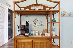 A coffee station with a sign reading 'But First... Coffee,' a Keurig coffee maker, and labeled containers for coffee, sugar, and tea. - Coastal Comfort Pet Friendly Barefoot Resort Villa (North Myrtle Beach)