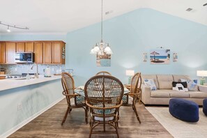 A modern kitchen with wooden cabinets, a microwave, and a gas stove. There is a dining area with wicker chairs and a table. A sofa and a blue ottoman are in the living room. - Coastal Comfort Pet Friendly Barefoot Resort Villa (North Myrtle Beach)