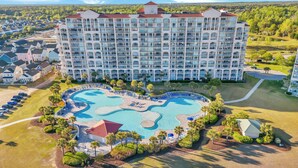 A large hotel with multiple balconies, a central swimming pool, and surrounding greenery. - Coastal Comfort Pet Friendly Barefoot Resort Villa (North Myrtle Beach)