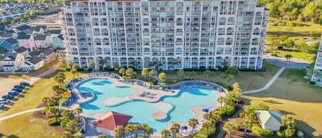 A large hotel with multiple balconies, a central swimming pool, and surrounding greenery.