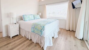 A bedroom with a bed, nightstand, lamp, window with blinds, and curtains. - Sandbag Castle 4bdr Waterfront (Orange Beach)