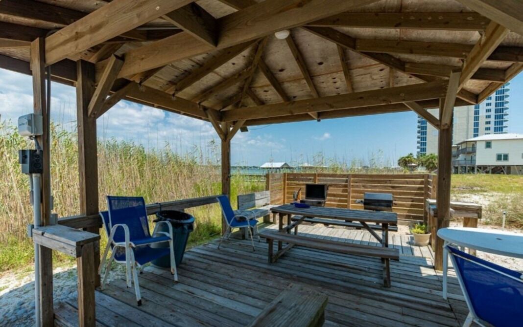 A wooden deck with picnic tables and chairs, overlooking a beach and a building in the distance.