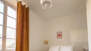 Un dormitorio con cama, una ventana con cortinas, una lámpara de pared y un cuadro pequeño.