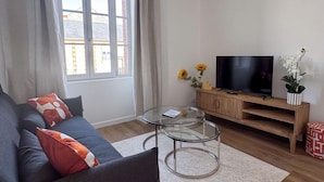 Una sala de estar con un sofá, una mesa de centro de vidrio, un televisor sobre un mueble de madera y una ventana con cortinas.