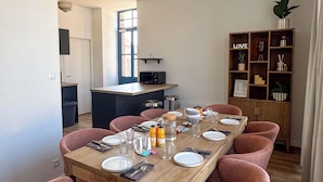 Comedor con una mesa de madera dispuesta para seis personas, rodeada de sillas rosas, y una cocina al fondo.