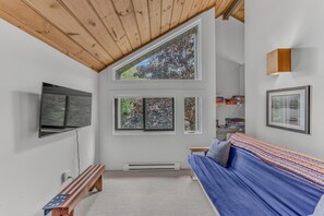 A room with a wooden ceiling, a blue sofa, a television mounted on the wall, and a window with a view of trees. - Modern Townhome at Loon- Pool & Hot Tub Access! (Lincoln)