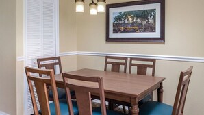 Un coin repas avec une table et des chaises en bois, un tableau encadré au mur et une fenêtre avec des volets blancs.