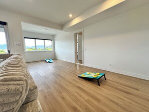 A spacious room with wooden flooring, a large window, and a door leading to another room. - The Malbec Residence: Live Luxury with your Favourite People! (West Kelowna)