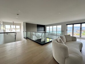 Ein modernes Wohnzimmer mit Balkon und Glasgeländer, einer Couch und Blick auf eine gebirgige Landschaft.