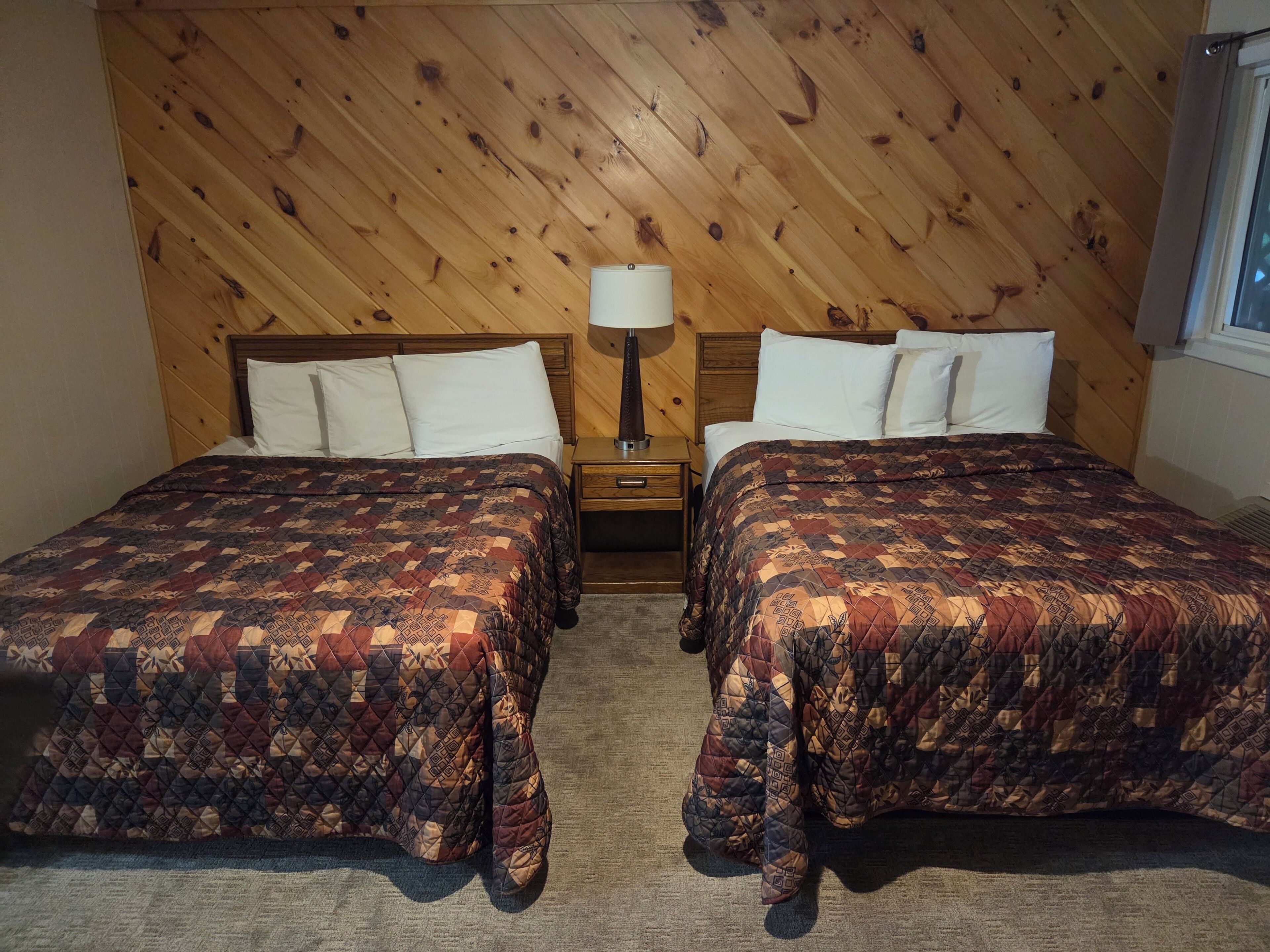 A room with two beds, a nightstand with a lamp, and a wooden wall.
