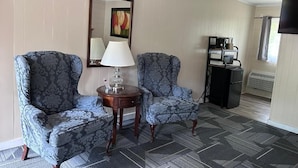 A hotel room with two patterned armchairs, a small table with a lamp, a painting on the wall, a coffee machine, and a TV mounted on the wall.