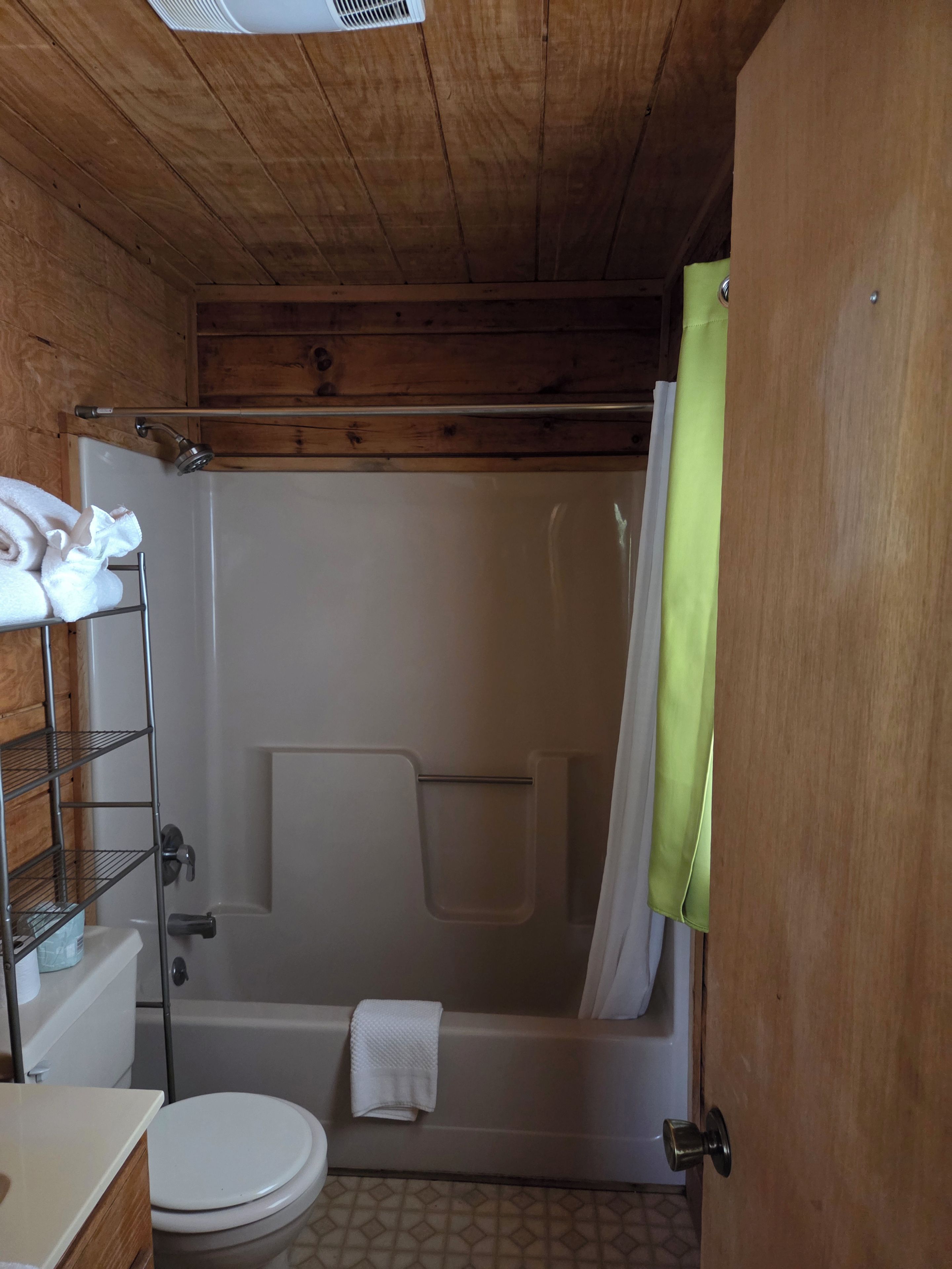 A wooden-paneled bathroom with a white toilet, a shower with a curtain, and a metal shelf with towels.