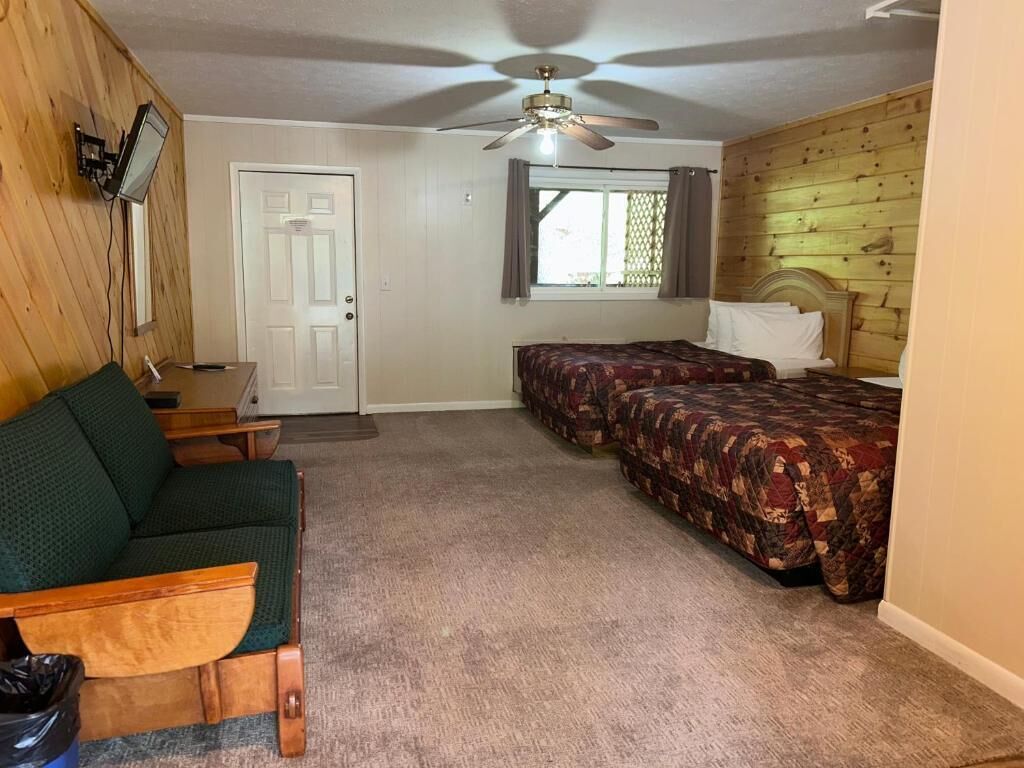 A room with a ceiling fan, two beds with patterned bedding, a wooden wall, a door, and a small table with a chair.
