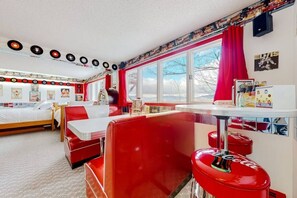 Ein stilvolles Zimmer im Retro-Design mit einem Bett, einer roten Sitzecke, einem Tisch mit einem Coca-Cola-Gasflasche und einem Fenster mit Aussicht.