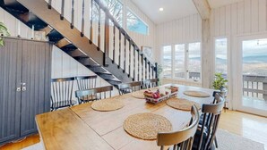Uma área de refeições com mesa de madeira, jogos americanos trançados e escada com corrimão preto.