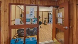 Quarto com painéis de madeira, equipamentos de ginástica, um ventilador e uma porta que dá para outra área.