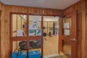 Ein Zimmer mit Holzwänden, Fitnessgeräten, einem Ventilator und einer Tür zu einem weiteren Bereich.