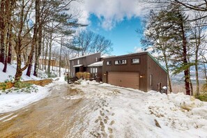 Un edificio de dos plantas con garaje, rodeado de nieve en el suelo y entre árboles.
