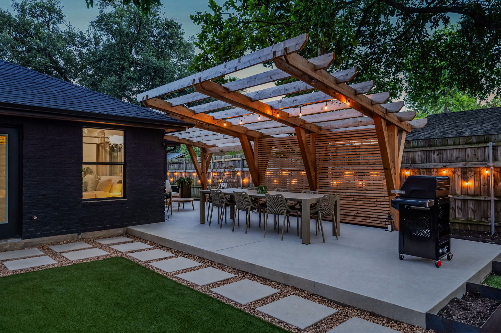 Une terrasse extérieure couverte, dotée d’une table et de chaises, d’un barbecue et d’une pergola illuminée de guirlandes lumineuses.