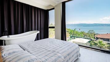 A bedroom with a large window, a bathtub, and a bed with striped bedding.