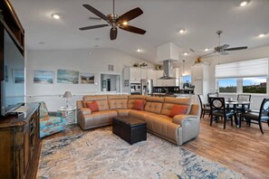 A spacious living room with a sectional sofa, a coffee table, and a dining area with a table and chairs. There are ceiling fans, recessed lighting, and a view of the outdoors through large windows. - Coastal Canary - Hot tub, beach, fire pit (Flagler Beach)