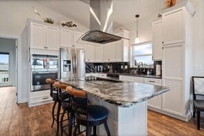 A modern kitchen with white cabinetry, a marble countertop, and a central island with bar stools. - Coastal Canary - Hot tub, beach, fire pit (Flagler Beach)