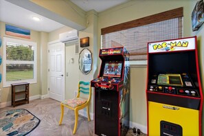 Ein Zimmer mit einem Pac-Man-Spielautomaten, einem Sessel, einem kleinen Tisch und einem Fenster mit Jalousien.