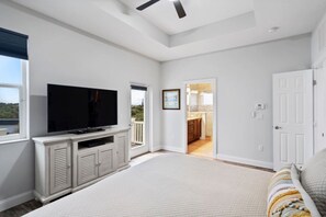 A bedroom with a TV, a door leading to a balcony, and a view of a kitchen. - Coastal Canary - Hot tub, beach, fire pit (Flagler Beach)