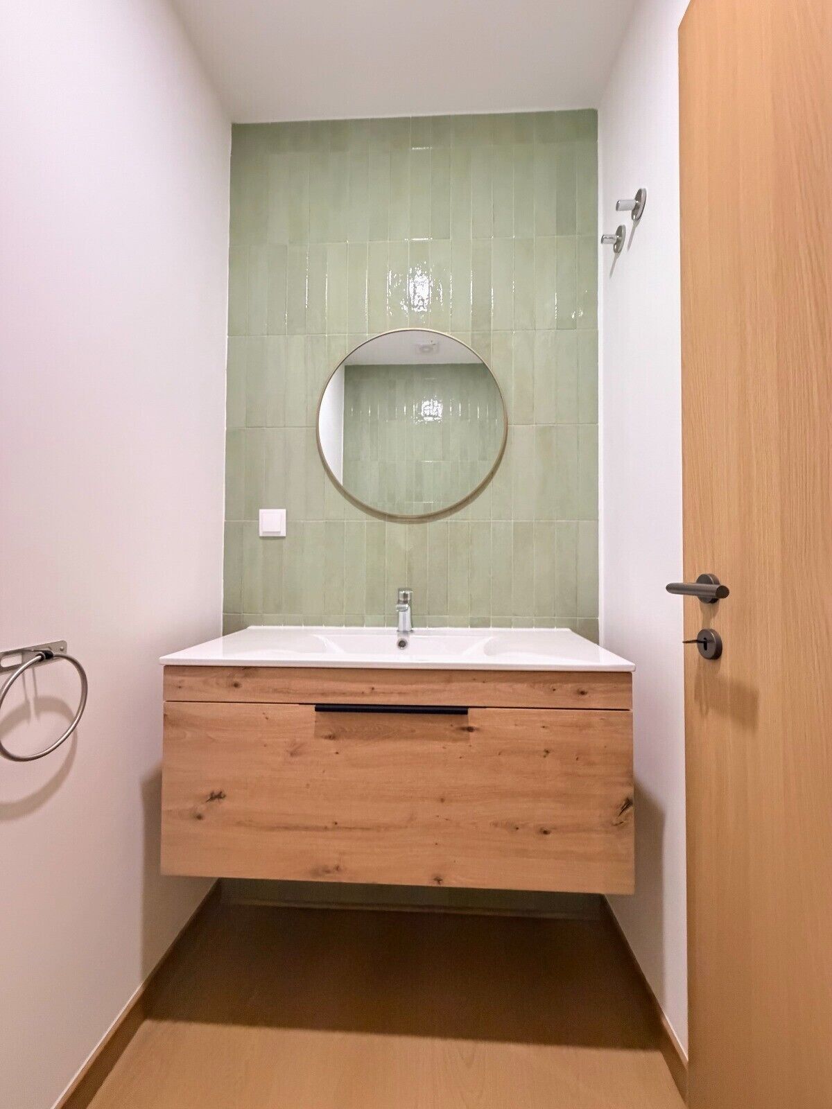 A bathroom with a wooden vanity, a round mirror, and a green-tiled wall.