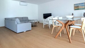 Una sala moderna con un sofá gris, una mesa de comedor con sillas blancas y un televisor de pantalla plana montado en la pared.