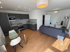 A modern kitchen with grey cabinets, a microwave, and a sink. There is a dining area with a glass table and chairs, and a grey sofa in the living area. - The Lyall - 1 Bedroom (London)